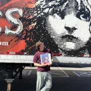 Person holding a Les Misérables booklet while standing in front of a large truck mural featuring the iconic Cosette illustration and red-and-black show artwork.