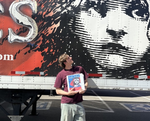 Person holding a Les Misérables booklet while standing in front of a large truck mural featuring the iconic Cosette illustration and red-and-black show artwork.
