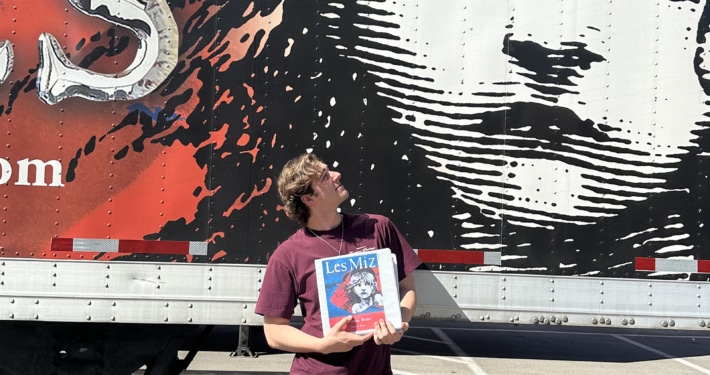 Person holding a Les Misérables booklet while standing in front of a large truck mural featuring the iconic Cosette illustration and red-and-black show artwork.