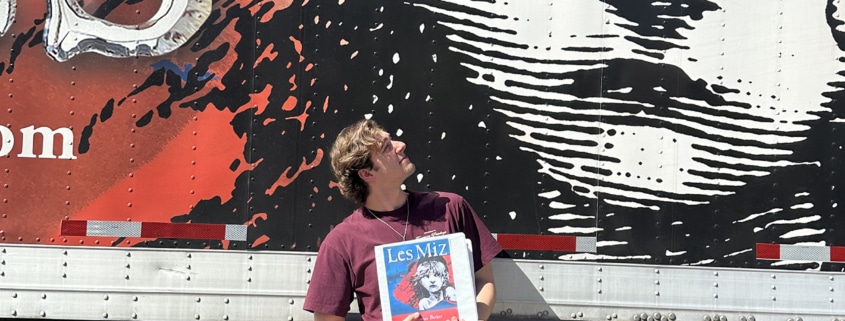 Person holding a Les Misérables booklet while standing in front of a large truck mural featuring the iconic Cosette illustration and red-and-black show artwork.