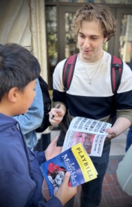 Person signing a Les Misérables Playbill for a young attendee outdoors, with the Playbill and autograph page visible in the foreground.