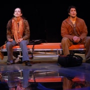 Two performers sit on an orange bench on a theater stage, with bags at their feet and a reflective floor beneath them under stage lighting.