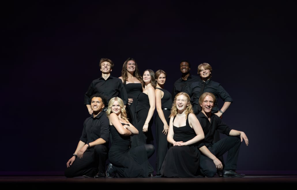 A group of ten stage performers dressed in coordinated black outfits pose together under a spotlight on a dark theater stage, arranged in two rows with some standing and others kneeling in the foreground.