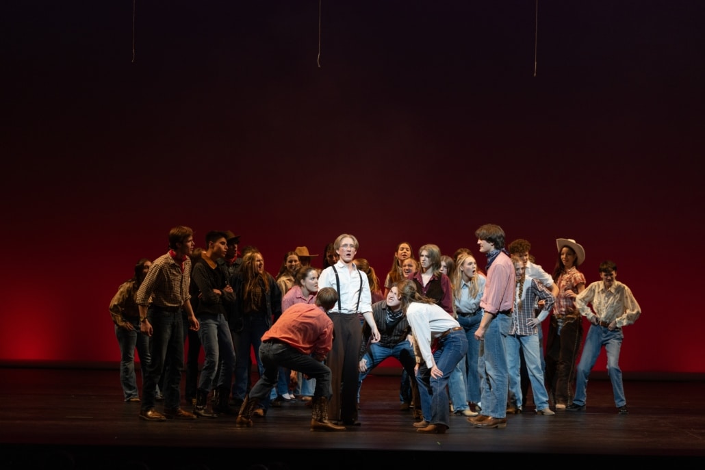 A large ensemble of stage performers in casual and western-inspired costumes gathers around a central figure in suspenders on a theater stage with a red-lit backdrop, with performers positioned in a semicircle and some leaning or crouching toward the center.
