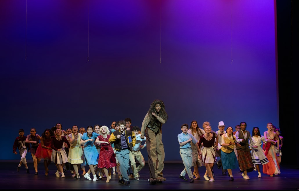 A wide group of performers in colorful costumes moves forward across a theater stage under blue and purple lighting, with one central figure in a textured costume and oversized feet leading the ensemble.