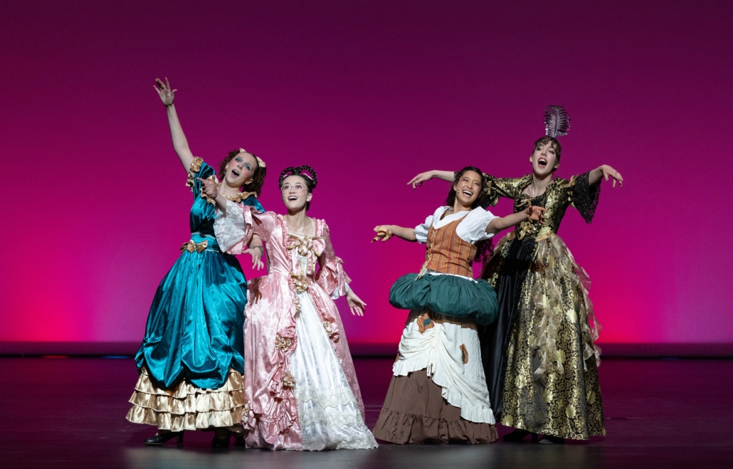 Four stage performers in colorful period costumes pose mid-song on a theater stage with a bright pink backdrop, arms extended outward in a coordinated musical number from Cinderella