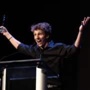 Performer in a black shirt stands at a podium on a dark stage holding an award and raising both arms while speaking into a microphone during an awards presentation.