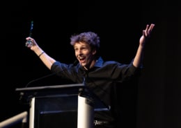 Performer in a black shirt stands at a podium on a dark stage holding an award and raising both arms while speaking into a microphone during an awards presentation.