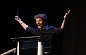 Performer in a black shirt stands at a podium on a dark stage holding an award and raising both arms while speaking into a microphone during an awards presentation.