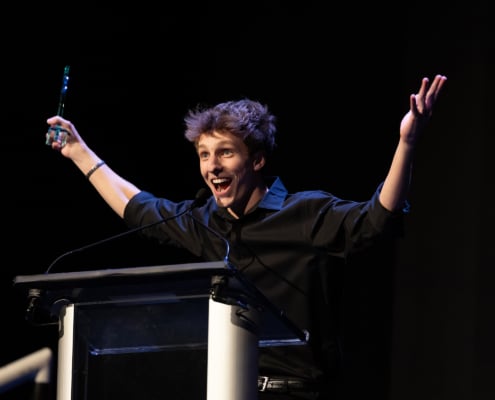 Performer in a black shirt stands at a podium on a dark stage holding an award and raising both arms while speaking into a microphone during an awards presentation.
