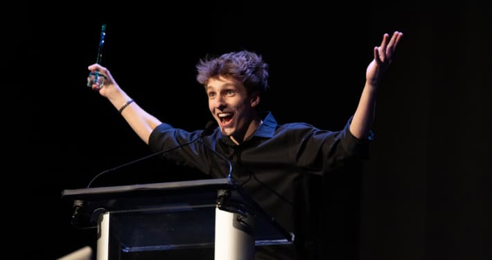 Performer in a black shirt stands at a podium on a dark stage holding an award and raising both arms while speaking into a microphone during an awards presentation.