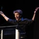 Performer in a black shirt stands at a podium on a dark stage holding an award and raising both arms while speaking into a microphone during an awards presentation.