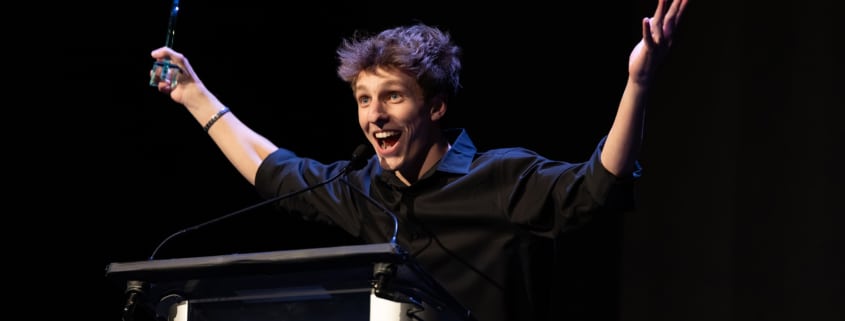 Performer in a black shirt stands at a podium on a dark stage holding an award and raising both arms while speaking into a microphone during an awards presentation.