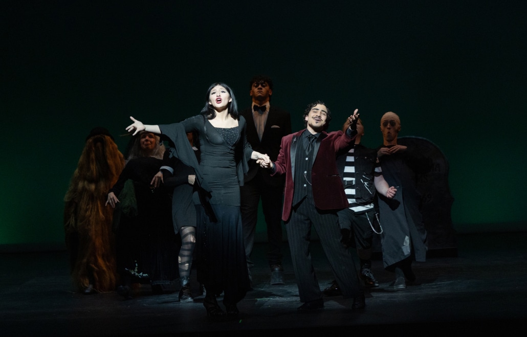 Stage scene from The Addams Family with two central performers in black costumes singing and holding hands while an ensemble in gothic-inspired outfits stands behind under dim green lighting.