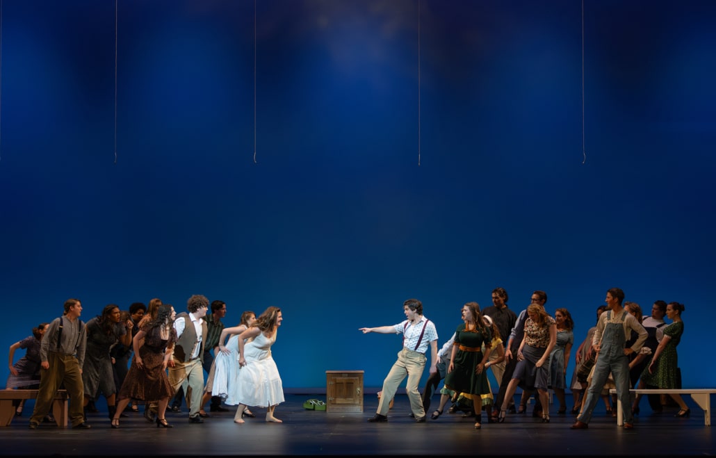 Large ensemble of performers in period-style costumes faces off across a stage with a blue backdrop as a central performer points during a dramatic moment in Bright Star.
