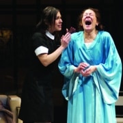 One performer in a black uniform stands beside another in a light blue robe who is leaning back with mouth open during an emotional moment on a stage set.