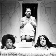 A performer stands centered behind a raised platform while two other performers’ heads appear at the front edge, surrounded by a rectangular structure resembling a display or stage set, presented in black-and-white lighting.