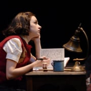 Performer seated at a small desk writing in a notebook, lit by a desk lamp on a dark stage with books and papers nearby.