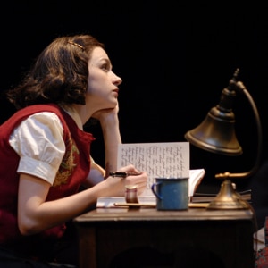 Performer seated at a small desk writing in a notebook, lit by a desk lamp on a dark stage with books and papers nearby.