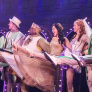 Cast members in colorful medieval costumes perform a high-kicking line dance on a stage set resembling a castle wall, with bright purple and pink theatrical lighting.