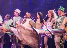 Cast members in colorful medieval costumes perform a high-kicking line dance on a stage set resembling a castle wall, with bright purple and pink theatrical lighting.