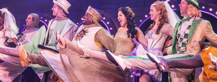 Cast members in colorful medieval costumes perform a high-kicking line dance on a stage set resembling a castle wall, with bright purple and pink theatrical lighting.