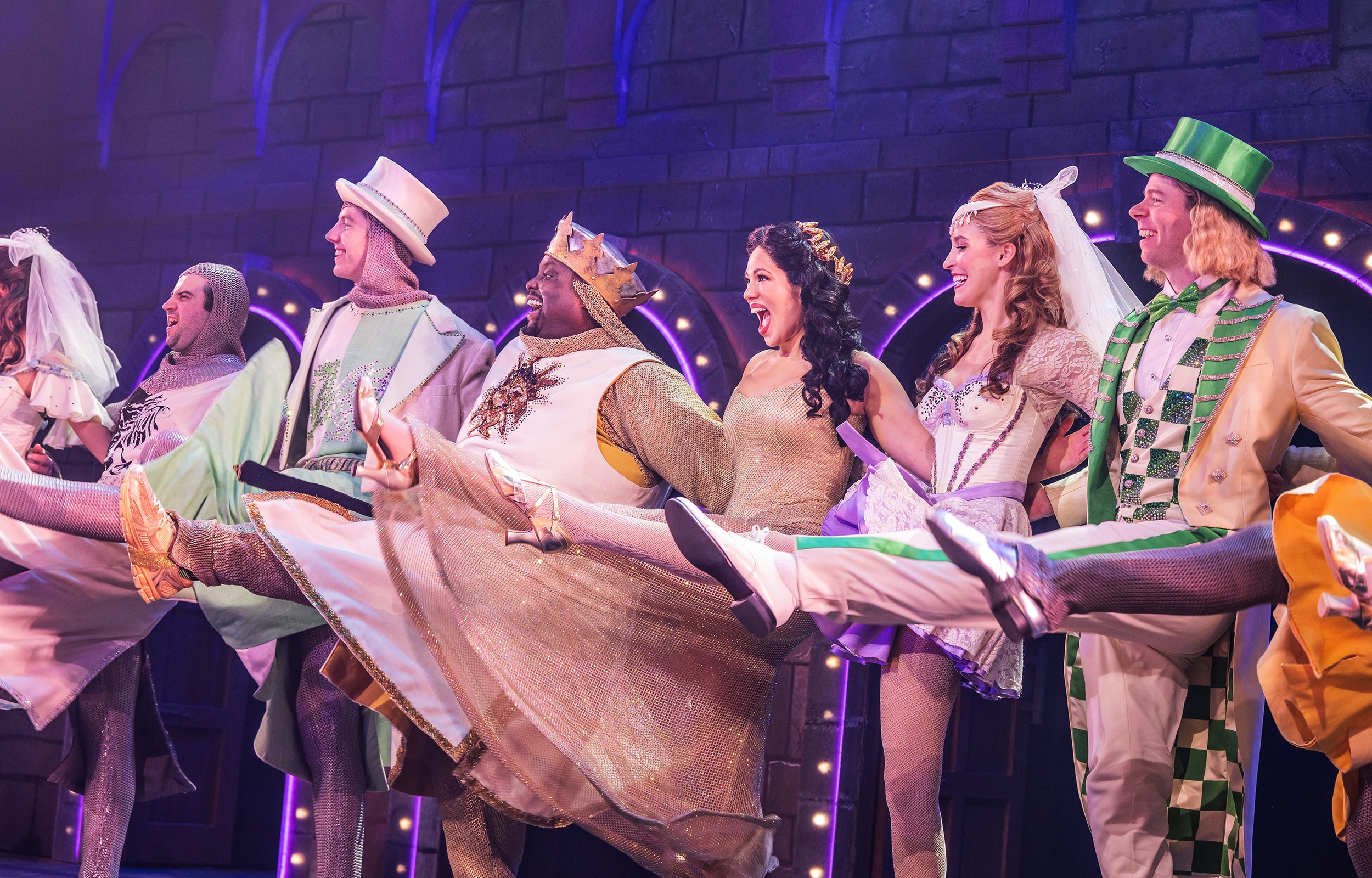 Cast members in colorful medieval costumes perform a high-kicking line dance on a stage set resembling a castle wall, with bright purple and pink theatrical lighting.