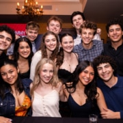 Large group of young people posing together at an indoor gathering, leaning in close and smiling under warm lighting with a chandelier overhead.