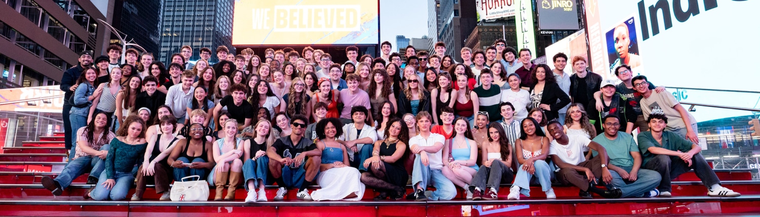 A very large group seated and standing on red steps in Times Square, surrounded by bright billboards and city skyscrapers, posing for a group photo.