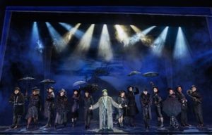 Full cast of the Beetlejuice North American tour stands in a line across the stage holding umbrellas, framed by dramatic spotlights and a dark, gothic backdrop with a twisted tree.