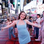 Person in a light blue dress standing on red steps in Times Square with arms outstretched, with a busy crowd and illuminated billboards in the background.