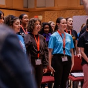 Group of young performers wearing name badges sing together inside a rehearsal room, standing in close formation with a conductor’s arm partially visible in the foreground.
