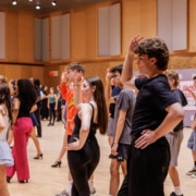 Large group of performers practicing choreography in a spacious rehearsal studio with wood floors, raising hands in unison during a dance sequence.