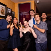 Small group of performers pose in a hallway pointing at a framed “Jimmy Awards” sign on the wall, standing in front of a gallery of framed photos.
