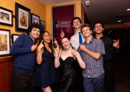 Small group of performers pose in a hallway pointing at a framed “Jimmy Awards” sign on the wall, standing in front of a gallery of framed photos.