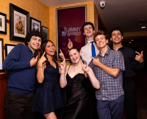 Small group of performers pose in a hallway pointing at a framed “Jimmy Awards” sign on the wall, standing in front of a gallery of framed photos.