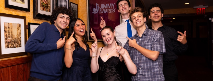 Small group of performers pose in a hallway pointing at a framed “Jimmy Awards” sign on the wall, standing in front of a gallery of framed photos.
