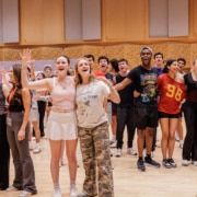 Large group of young performers singing together in a rehearsal room, some with arms around each other as they face forward.