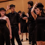 Performers in black outfits rehearse choreography in a studio, lifting bowler hats in a synchronized gesture on a wooden floor.