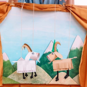 Handmade puppet theater scene with two hanging paper goats against a painted mountain backdrop, framed by orange curtains.