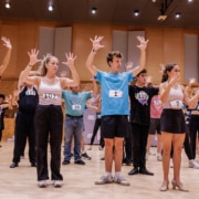 Performers wearing numbered audition tags practice choreography in rows, raising both hands in unison inside a rehearsal studio.