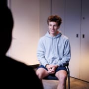 Person sitting on a chair in a softly lit room during an interview, with a camera setup visible at the side and another person partially visible in the foreground.