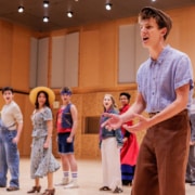 Performer in a flat cap and vintage-style costume gestures while rehearsing a scene, with a line of other performers in colorful outfits standing behind in a studio space.