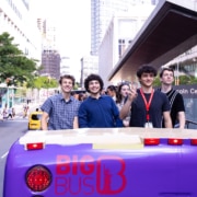 Group of young people standing on the back of a purple open-top bus in a city street near Lincoln Center Theater, with buildings and pedestrians in the background.