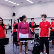 Group of performers in red shirts standing at music stands during a rehearsal in a studio, singing together in a circle with mirrored walls in the background.
