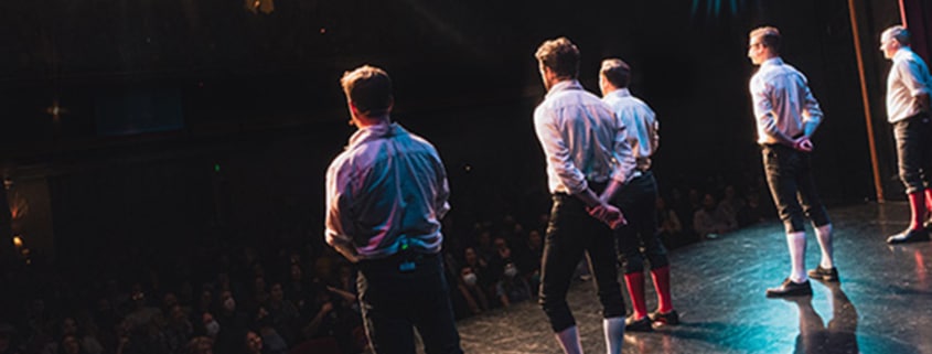 Performers in period-style costumes standing on a theater stage facing a seated audience under dramatic stage lighting.