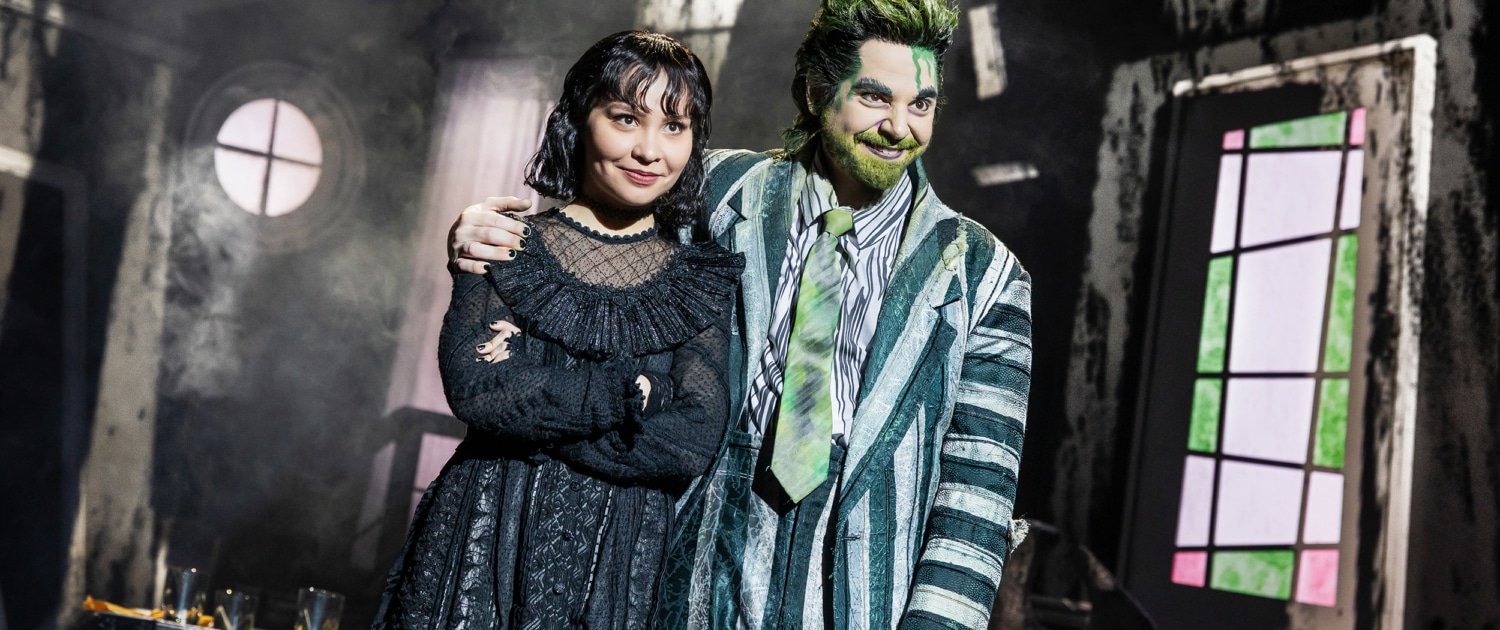 Two performers stand close together in a distressed interior set, one in a striped suit with green accents and one in a black gothic outfit, framed by a round window and angled walls.