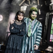 Two performers stand close together in a distressed interior set, one in a striped suit with green accents and one in a black gothic outfit, framed by a round window and angled walls.