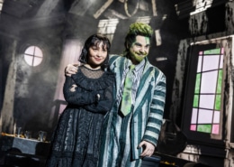 Two performers stand close together in a distressed interior set, one in a striped suit with green accents and one in a black gothic outfit, framed by a round window and angled walls.