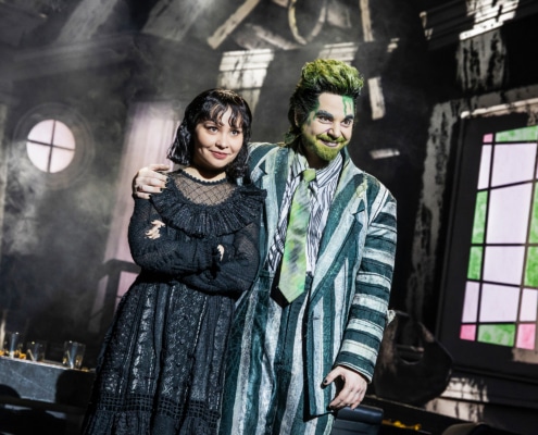 Two performers stand close together in a distressed interior set, one in a striped suit with green accents and one in a black gothic outfit, framed by a round window and angled walls.